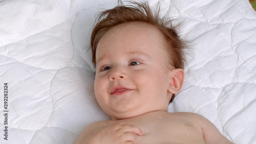 White smiling little child with dark thick hair and dark eyes is lying topless on the blanket on open air and observes the area. It is moving head and fingers. It raises left and right hand White smiling little child with dark thick hair and dark eyes is lying topless on the blanket on open air and observes the area. It is moving head and fingers. It raises left and right hand