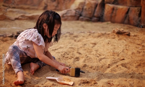 A young Asian girl playing in a sandbox with a modeled dinosaur fossil using brush and shovel.