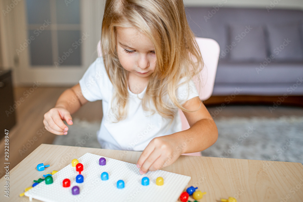 Little blonde girl in a white tshirt playing with plastic multicolor mosaic at home or preschool. Early education concept. Little blonde girl in a white tshirt playing with plastic multicolor mosaic at home or preschool. Early education concept.