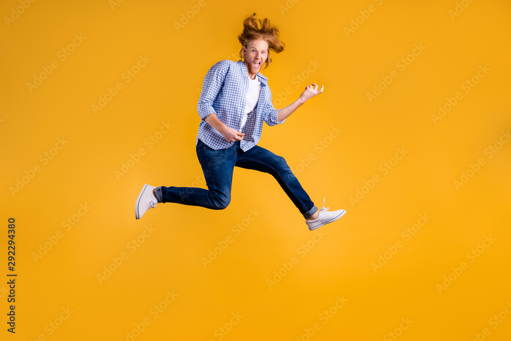 Full length body size photo of crazy funny red haired rock hard rocker man fan wearing jeans denim checkered blue shirt sneakers pretending to play guitar jumping isolated vivid color background