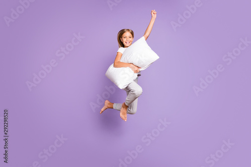 Full length photo of positive cheerful funny kid jump raise fist ahead imagine she strong superhero fly with pillow wear white comfort t-shirt pants barefoot isolated violet purple color background