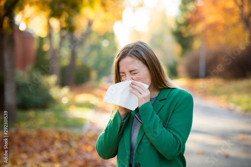 Sick young woman with cold and flu standing outdoors, sneezing, wiping nose with handkerchief, coughing. Autumn street background