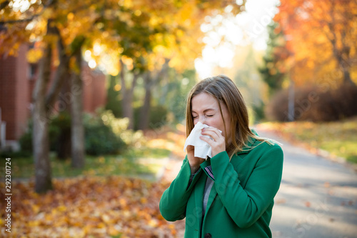 Sick young woman with cold and flu standing outdoors, sneezing, wiping nose with handkerchief, coughing. Autumn street background photo