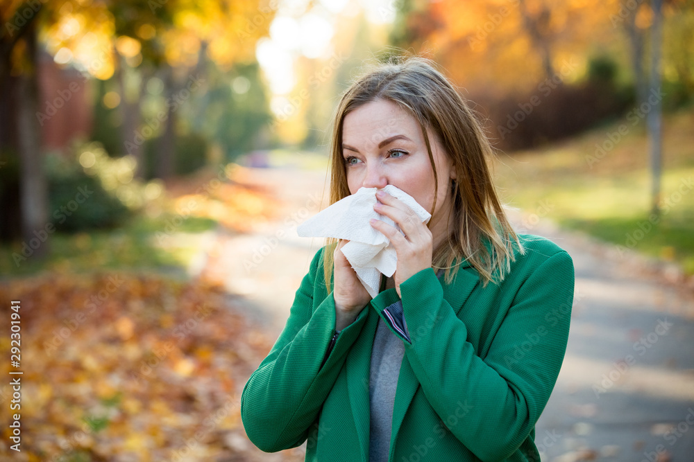 Sick young woman with cold and flu standing outdoors, sneezing, wiping nose with handkerchief, coughing. Autumn street background Sick young woman with cold and flu standing outdoors, sneezing, wiping nose with handkerchief, coughing. Autumn street background