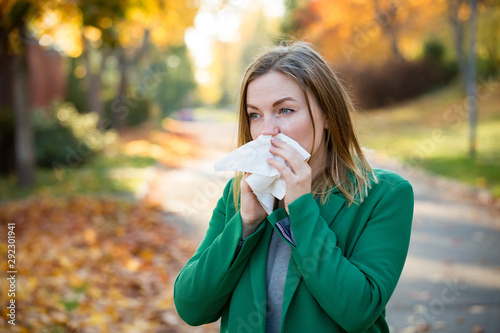 Sick young woman with cold and flu standing outdoors, sneezing, wiping nose with handkerchief, coughing. Autumn street background