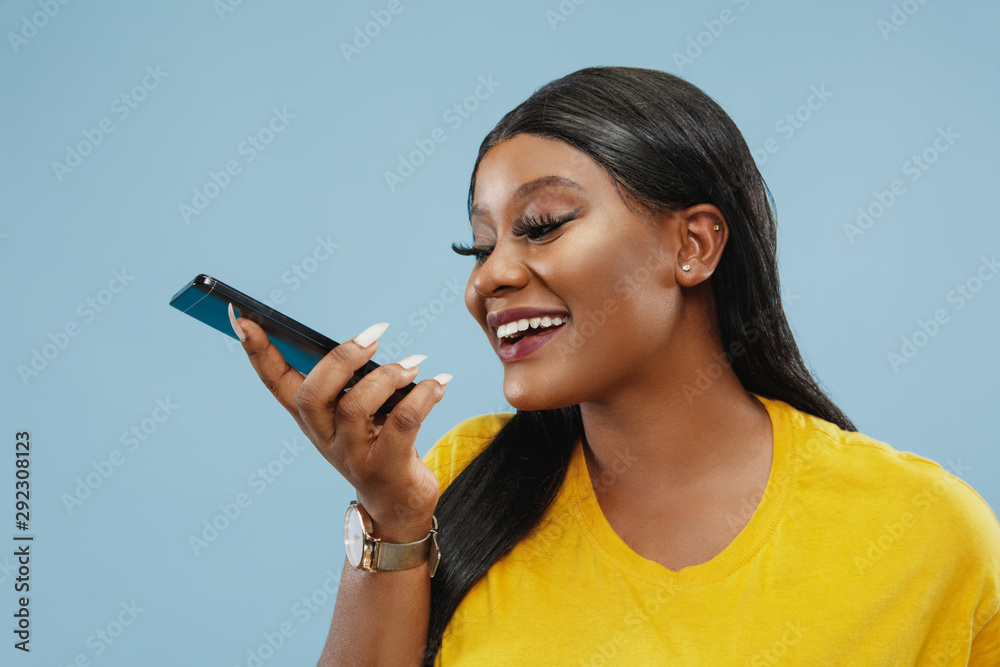 African-american young woman's half-length portrait on blue studio background. Beautiful female model. Concept of human emotions, facial expression, sales, ad. Recording voice message. African-american young woman's half-length portrait on blue studio background. Beautiful female model. Concept of human emotions, facial expression, sales, ad. Recording voice message.