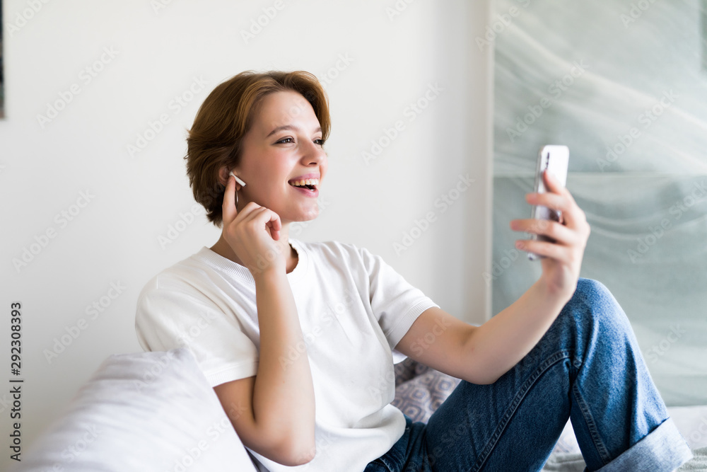 Smiling girl relaxing at home playing music using a smartphone and wearing white headphones Smiling girl relaxing at home playing music using a smartphone and wearing white headphones
