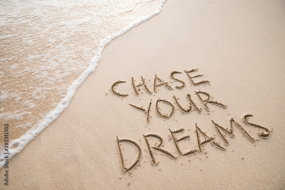 Fototapeta premium Chase Your Dreams travel message handwritten on smooth sand beach with incoming tropical wave