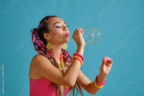 Lovely girl with a multi-colored braids hairstyle and bright make-up, is blowing bubbles using her hands, posing in studio against a blue background.
