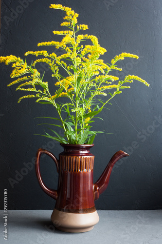 bouquet of canadian goldenrod in a brown ceramic teapot on a gray background, twigs with delicate yellow flowers