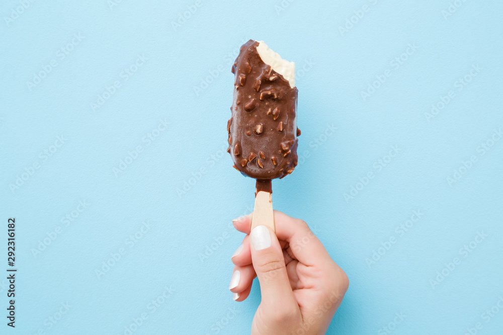 Young woman hand holding white vanilla ice cream with nuts and chocolate glaze on pastel light blue background. Bitten food. Closeup. - obrazy, fototapety, plakaty 