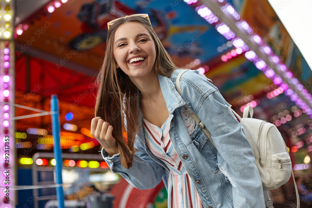 Cheerful young pretty woman with long brown hair walking in park of attractions, wearing casual clothes and white backpack, holding her hair and looking to camera with wide sincere smile Cheerful young pretty woman with long brown hair walking in park of attractions, wearing casual clothes and white backpack, holding her hair and looking to camera with wide sincere smile