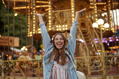 Happy young brunette lady with long hair standing over carousel in park of attractions, raising hands joyfully with wide mouth opened, positive emotions concept