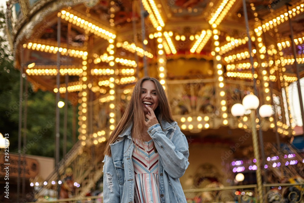 Attractive young long haired brunette lady walking through amusement park in romantic dress and jeans coat, looking to camera with broad sincere smile and keeping forefinger on her underlip