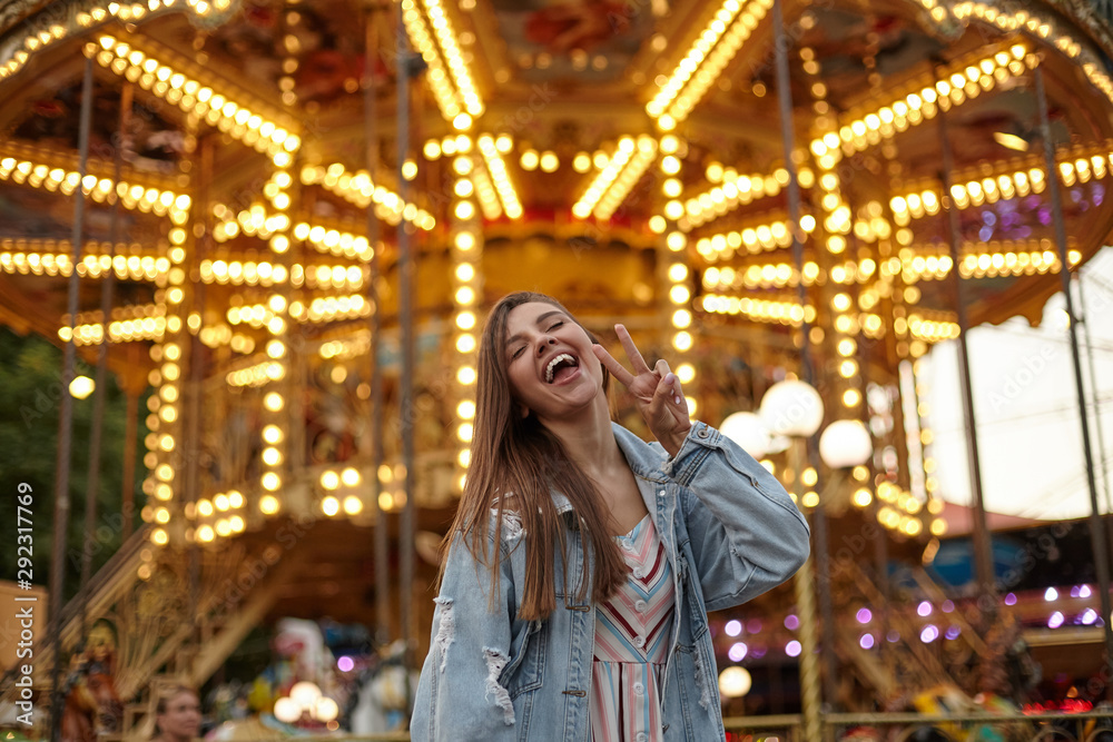Joyful young attractive woman with brown long hair posing over carousel in jeans coat, raising hand with victory gesture, smiling with wide mouth opened and closing eyes