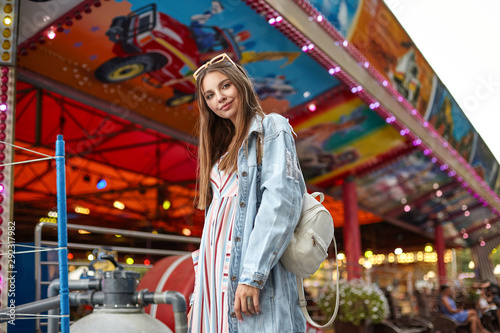 Beautiful young long haired female with sunglasses on her head posing over attractions in amusement park, wearing romantic summer dress and trendy jeans coat