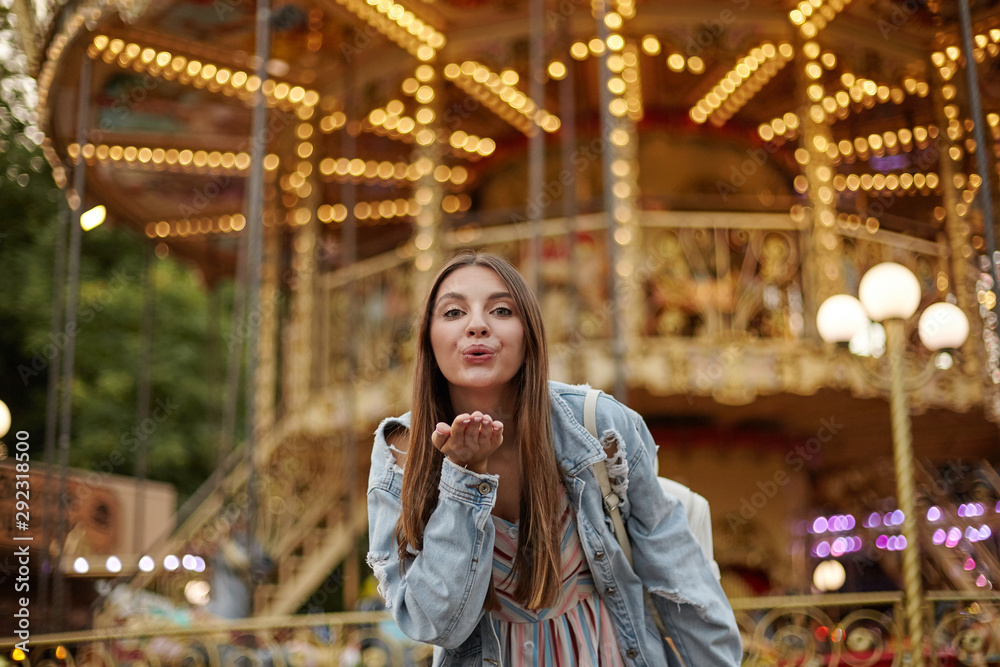 Outdoor photo of charming long haired young lady in trendy jeans coat raising palm and blowing air kiss to camera, standing over carousel in park of attractions Outdoor photo of charming long haired young lady in trendy jeans coat raising palm and blowing air kiss to camera, standing over carousel in park of attractions