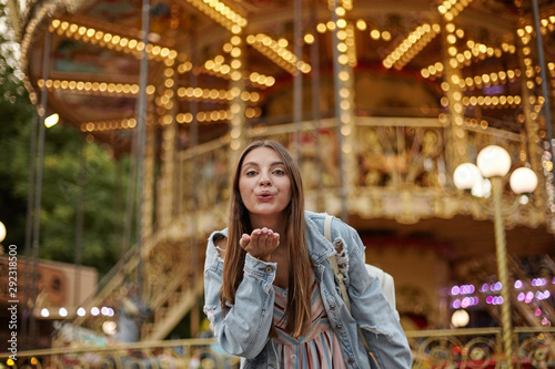 Outdoor photo of charming long haired young lady in trendy jeans coat raising palm and blowing air kiss to camera, standing over carousel in park of attractions © timtimphoto