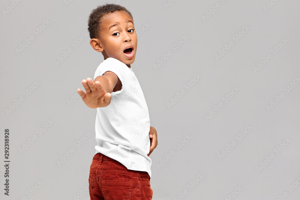 Body language. Isolated shot of funny dark skinned boy in white t-shirt and corduroy trousers outstretching arm, making stop gesture, exclaming No, having scared or shocked facial expression Body language. Isolated shot of funny dark skinned boy in white t-shirt and corduroy trousers outstretching arm, making stop gesture, exclaming No, having scared or shocked facial expression