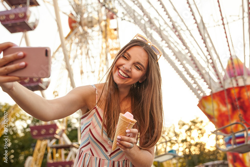 Beautiful young long haired brunette lady in romantic summer dress posing over attractions while making selfie with her mobile phone, keeping ice cream cone in hand and smiling cheerfully to camera