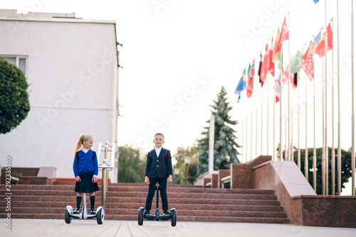 Two young schoolchildren  riding on gyroscooters.  Kids go to school for gyroscooters. Blonde kids. Back to school. Modern schoolchildren. Kids in school uniform. Flags of foreign countries. photo