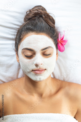 Nice relaxing young woman with white mask on face lying down on towel on bench spa with flower on hair. Smiling on camera.