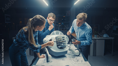 Late at Night in Robotics Engineering Facility Three Technical Engineers Talk and Work on a Wheeled Robot Prototype. In the Background High Tech Research Center with Screens Showing Industrial Design