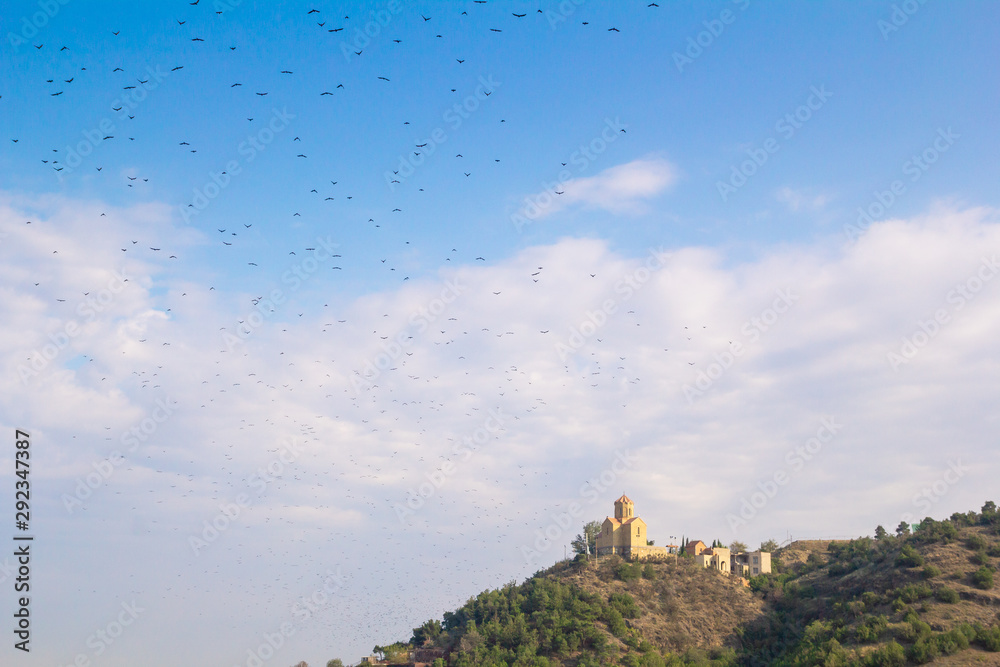 View of the church building standing on a hill. A large flock of small birds on a blue summer sky with white clouds