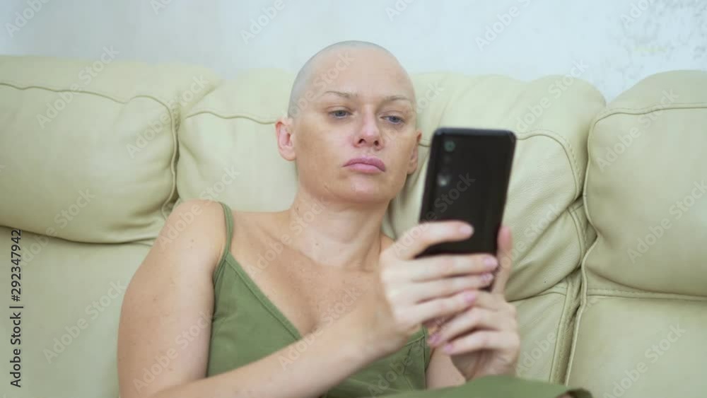 bald woman uses a smartphone while sitting on a sofa bald woman uses a smartphone while sitting on a sofa