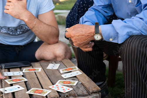 Close up photo of two men, younger in casual clothes, older in elegant clothes that playing cards on a wooden box outdoor