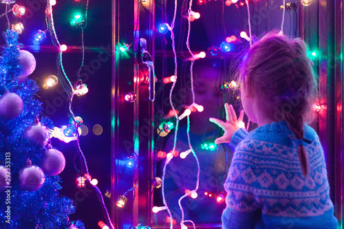 Happy little cute, beautiful girl 5 years old is sitting on the window and playing with Christmas lights and  Christmas decoration. Dark-blue theme. 