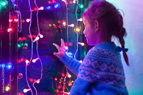 Happy little cute, beautiful girl 5 years old is sitting on the window and playing with Christmas lights and  Christmas decoration. Dark-blue theme. 