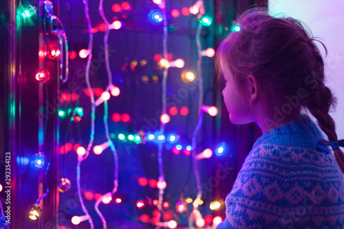 Happy little cute, beautiful girl 5 years old is sitting on the window and playing with Christmas lights and  Christmas decoration. Dark-blue theme. 