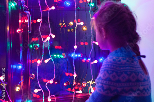 Happy little cute, beautiful girl 5 years old is sitting on the window and playing with Christmas lights and  Christmas decoration. Dark-blue theme. 