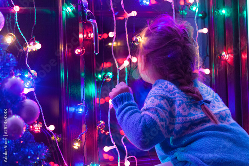 Happy little cute, beautiful girl 5 years old is sitting on the window and playing with Christmas lights and  Christmas decoration. Dark-blue theme. 