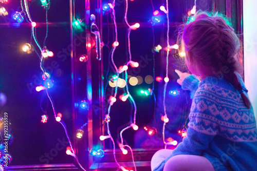 Happy little cute, beautiful girl 5 years old is sitting on the window and playing with Christmas lights and  Christmas decoration. Dark-blue theme. 