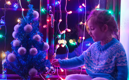 Happy little cute, beautiful girl 5 years old is sitting on the window and playing with Christmas lights and  Christmas decoration. Dark-blue theme. 
