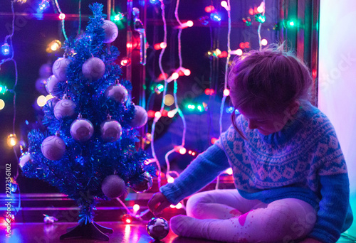 Happy little cute, beautiful girl 5 years old is sitting on the window and playing with Christmas lights and  Christmas decoration. Dark-blue theme. 