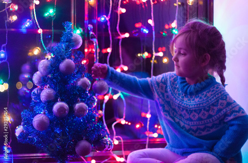 Happy little cute, beautiful girl 5 years old is sitting on the window and playing with Christmas lights and  Christmas decoration. Dark-blue theme. 