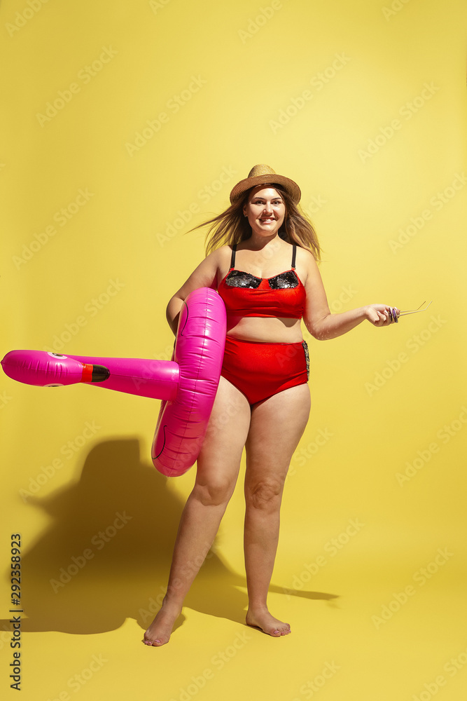 Young caucasian plus size female model's preparing for beach resort on yellow background. Woman in red swimsuit, hat and sunglasses. Concept of summertime, party, body positive. Swimring as a flamingo