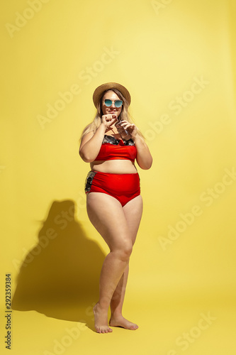 Young caucasian plus size female model's preparing for beach resort on yellow background. Woman in red swimsuit, hat and sunglasses. Concept of summertime, party, body positive. Drinking cocktail.
