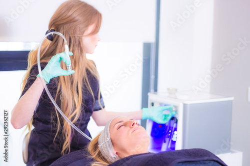Woman lying and relaxed in an aesthetic medicine clinic while the beautician adjusts the parameters of the high-tech machine for facial treatment