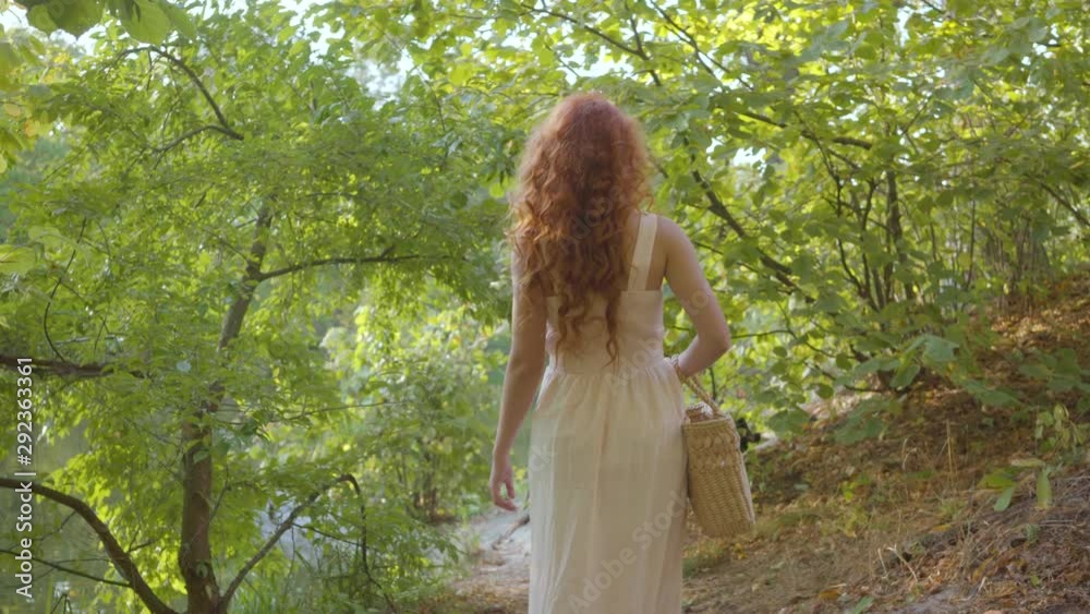 Back view of a young nice woman dressed in light white clothes walking in the autumn forest with a straw basket. Attractive fairytale girl turning to the camera and smiling mysteriously. Back view of a young nice woman dressed in light white clothes walking in the autumn forest with a straw basket. Attractive fairytale girl turning to the camera and smiling mysteriously.