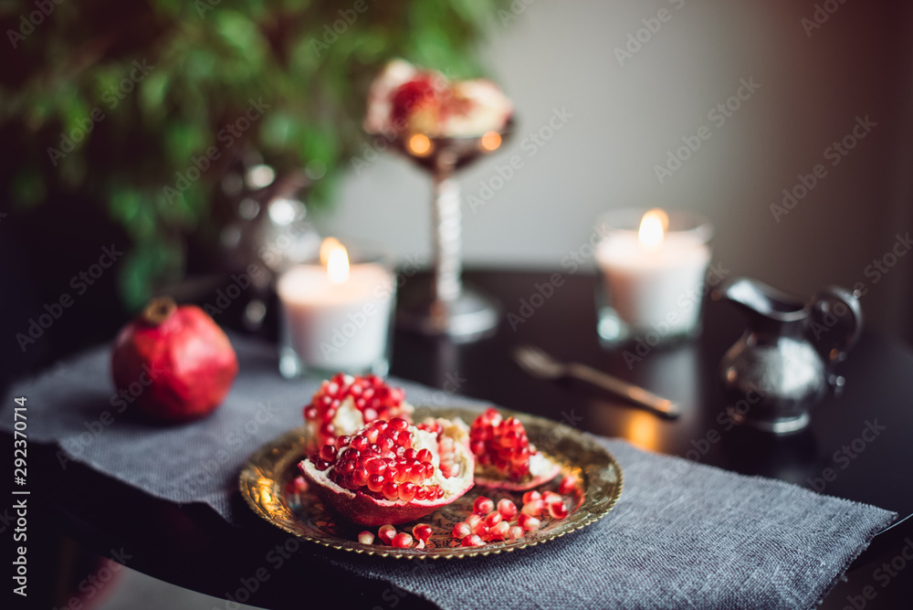 Still life composition with juicy red cut cleared pomegranate on a copper plate, burning candles and other oriental decor on the black table. Soft selective focus. Copy space.