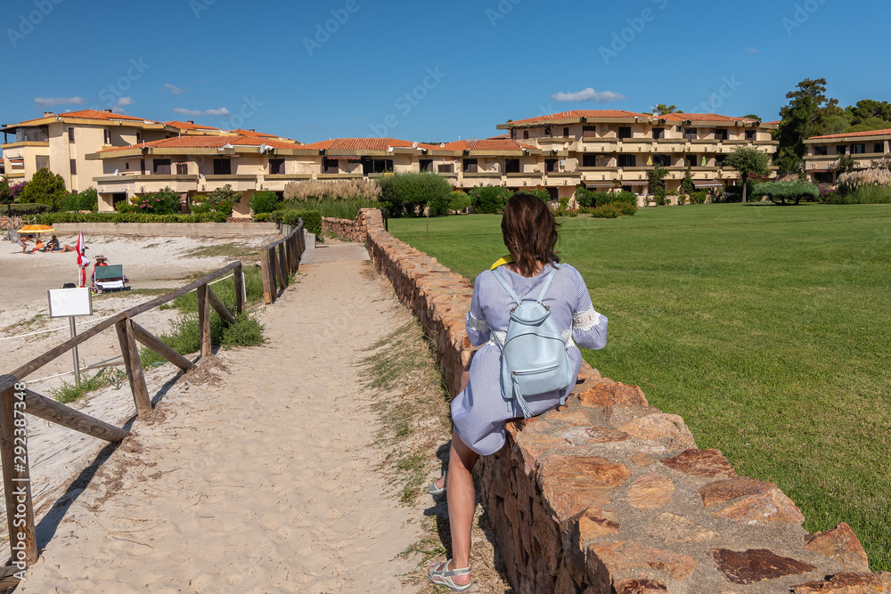 Woman tourist with a backpack sits on a stone fence and looks at the lawn and cottages