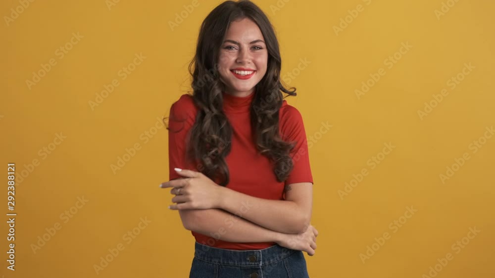 Pretty flirty brunette girl in red top with hands crossed happily lightly dancing on camera over yellow background
