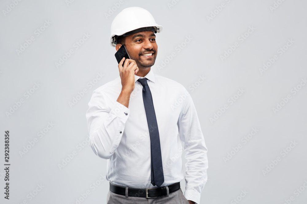 architecture, construction business and people concept - smiling indian male architect in helmet calling on smartphone over grey background