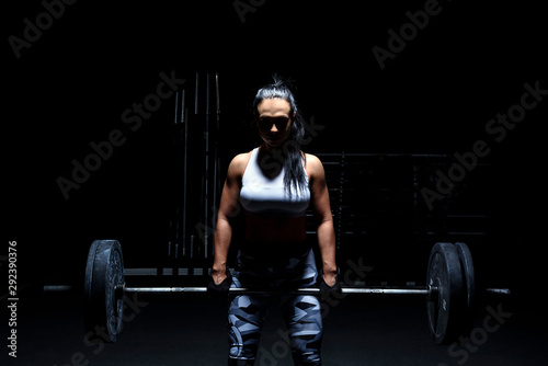 Woman bodybuilder make her workout with weights - barbell. Light from above, dark background, mysterious photo.