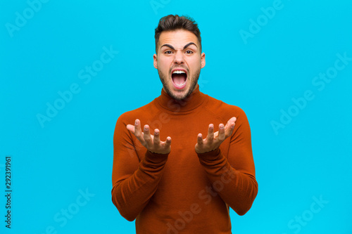 young hispanic man looking desperate and frustrated, stressed, unhappy and annoyed, shouting and screaming against blue background