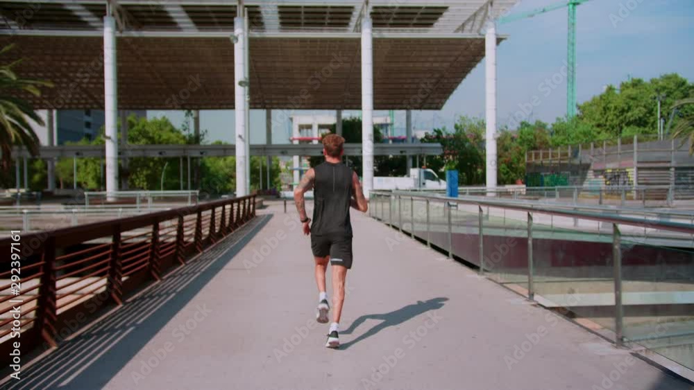 Back side shoot of strong man in black sportswear running at sunny morning urban bridge, sport goals every day at summer leisure motivation of tall male in fit body shape with sun panel on background Back side shoot of strong man in black sportswear running at sunny morning urban bridge, sport goals every day at summer leisure motivation of tall male in fit body shape with sun panel on background